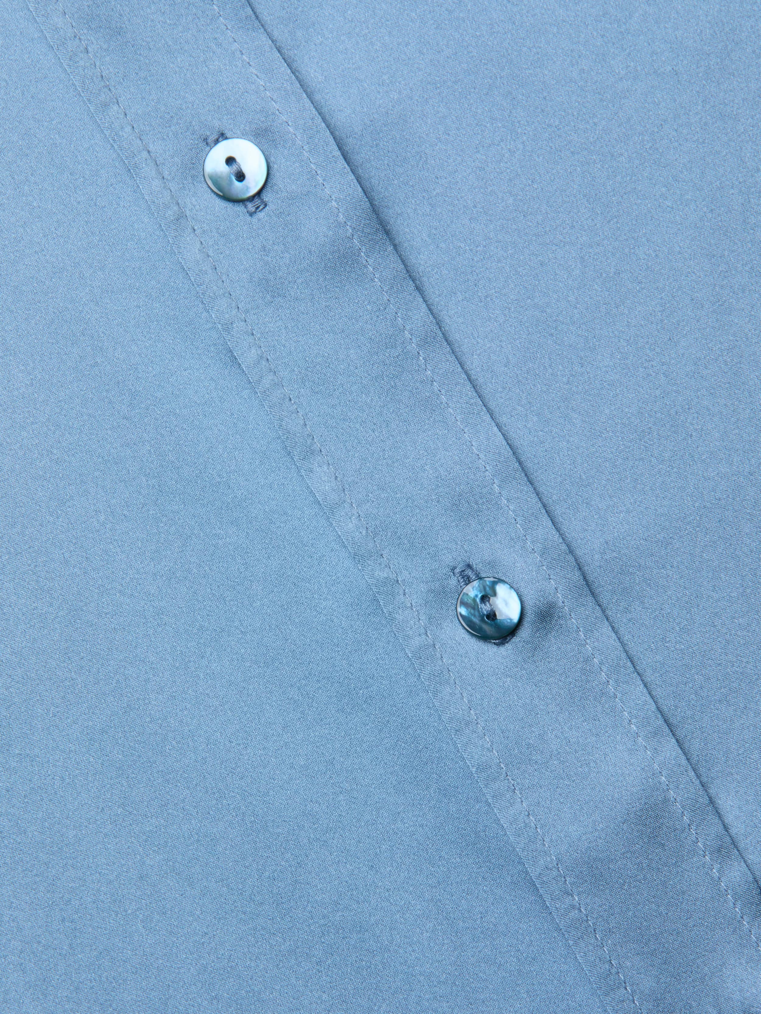 Close-up of a blue mist Dani Silk Blouse fabric with two small, shiny buttons along a stitched seam, shown at a slight diagonal angle.