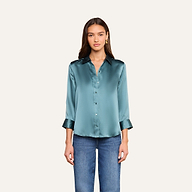 Una mujer posa frente a un fondo liso de color beige, mirando hacia delante con expresión neutra, vestida con la blusa de seda «Dani» en azul cadete brillante —con botones en la parte delantera y mangas tres cuartos— combinada con unos vaqueros azules.