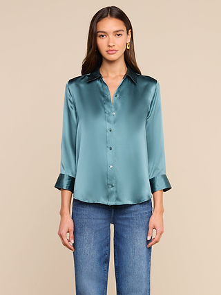 A woman stands against a plain beige background, facing forward with a neutral expression, wearing the Dani Silk Blouse in shiny cadet blue—featuring a button-down front and 3/4 sleeves—paired with blue jeans.