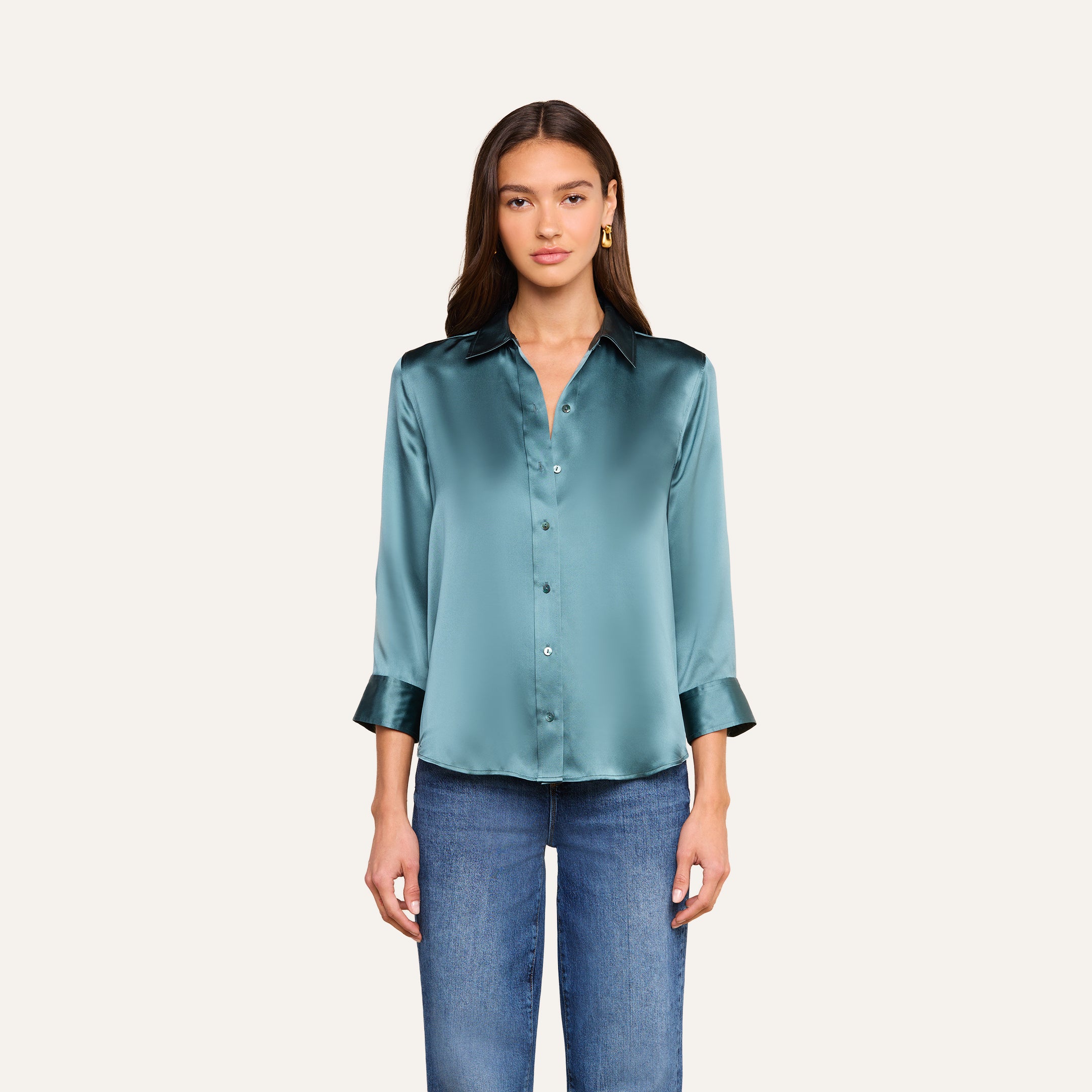 Una mujer posa frente a un fondo liso de color beige, mirando hacia delante con expresión neutra, vestida con la blusa de seda «Dani» en azul cadete brillante —con botones en la parte delantera y mangas tres cuartos— combinada con unos vaqueros azules.