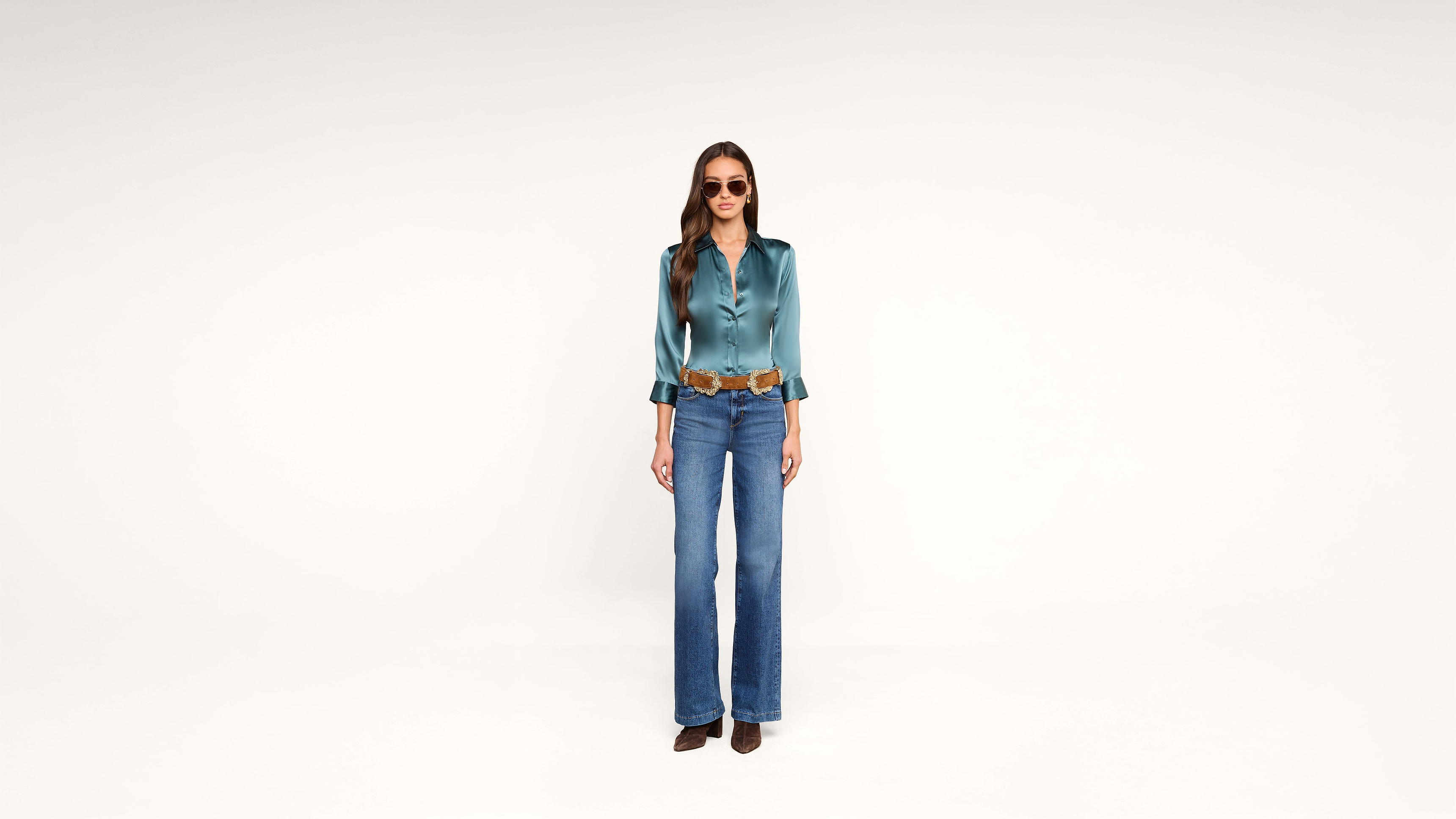 Woman stands against a plain white background wearing sunglasses, a teal satin blouse, a brown belt, wide-leg blue jeans, and brown shoes. She has long brown hair styled in loose waves.