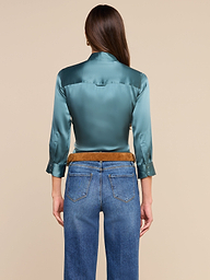 Una mujer con el pelo largo y castaño, de espaldas a la cámara y con un fondo beige, lleva una blusa brillante de Dani Silk en color verde azulado con mangas tres cuartos, combinada con unos vaqueros azules y un cinturón marrón.