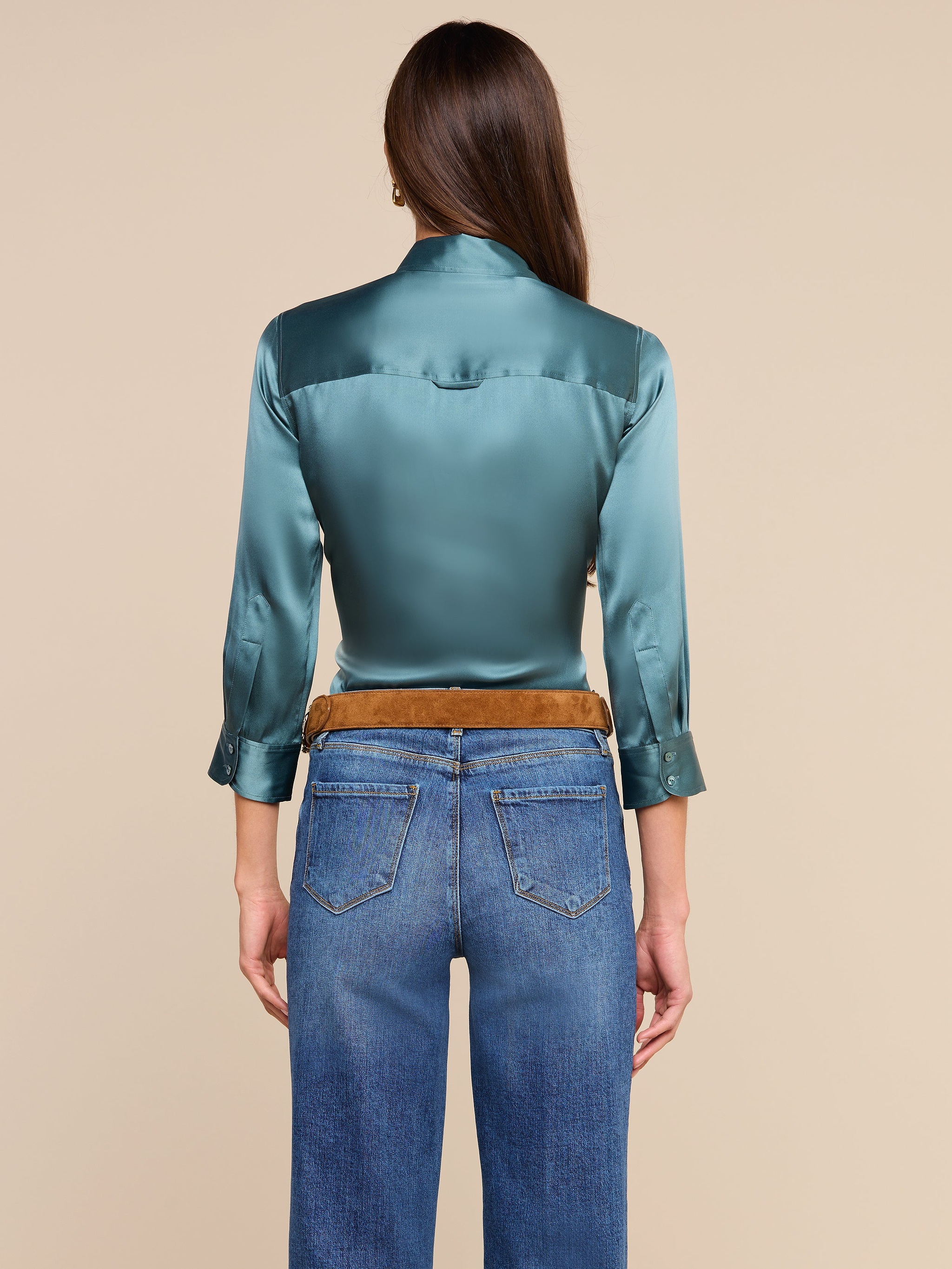 Una mujer con el pelo largo y castaño, de espaldas a la cámara y con un fondo beige, lleva una blusa brillante de Dani Silk en color verde azulado con mangas tres cuartos, combinada con unos vaqueros azules y un cinturón marrón.