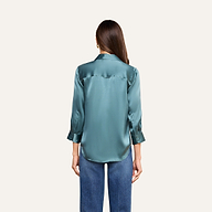 Una mujer con el pelo largo y castaño luce la blusa de seda «Dani» en azul cadete —una lujosa blusa de seda con botones y puños enrollados— combinada con unos vaqueros azules, de pie de espaldas a la cámara sobre un fondo beige.