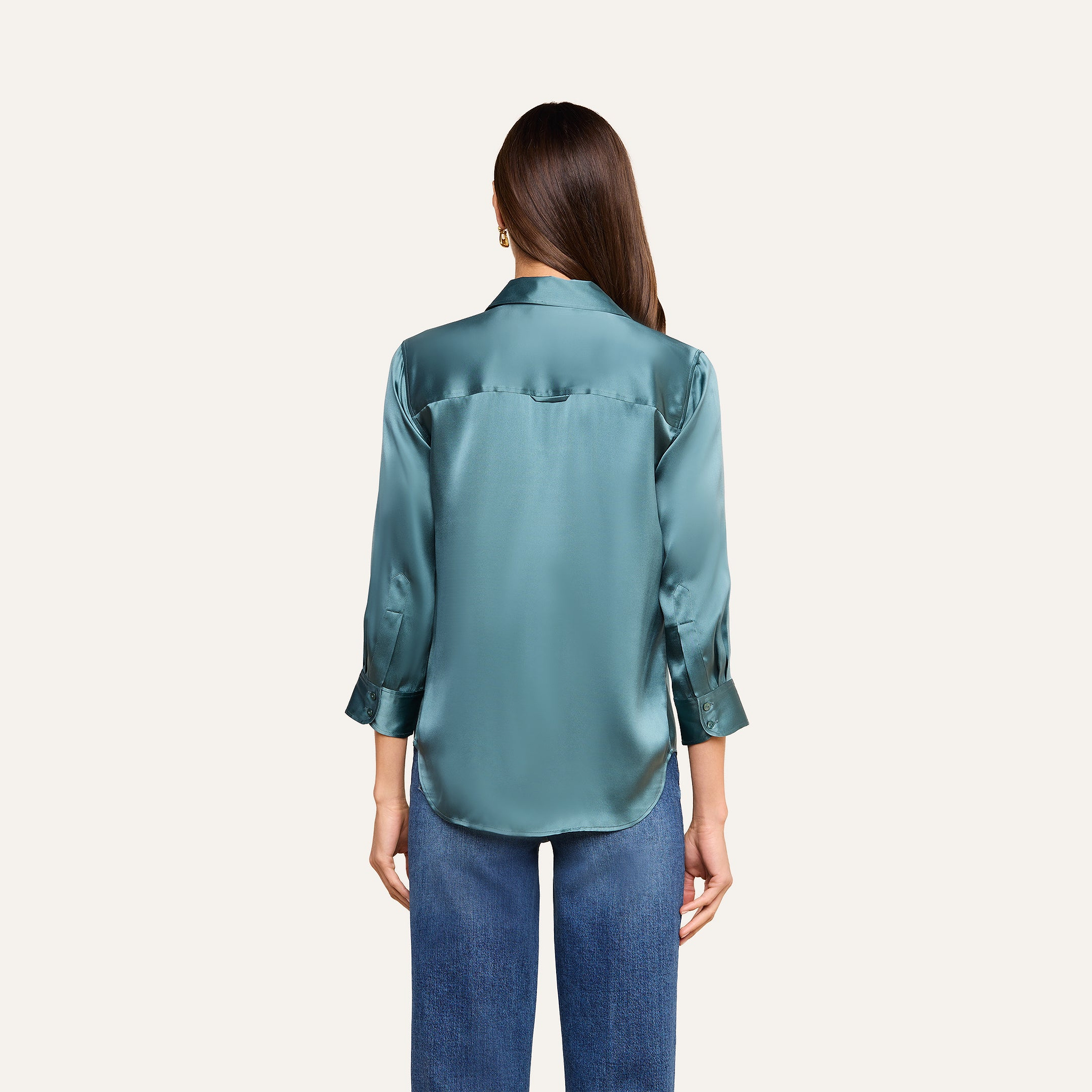 Una mujer con el pelo largo y castaño luce la blusa de seda «Dani» en azul cadete —una lujosa blusa de seda con botones y puños enrollados— combinada con unos vaqueros azules, de pie de espaldas a la cámara sobre un fondo beige.