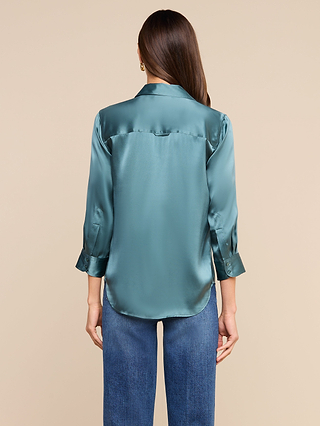 A woman with long brown hair models the Dani Silk Blouse in cadet blue—a luxurious silk button-down featuring rolled cuffs—paired with blue jeans, standing with her back to the camera against a beige background.
