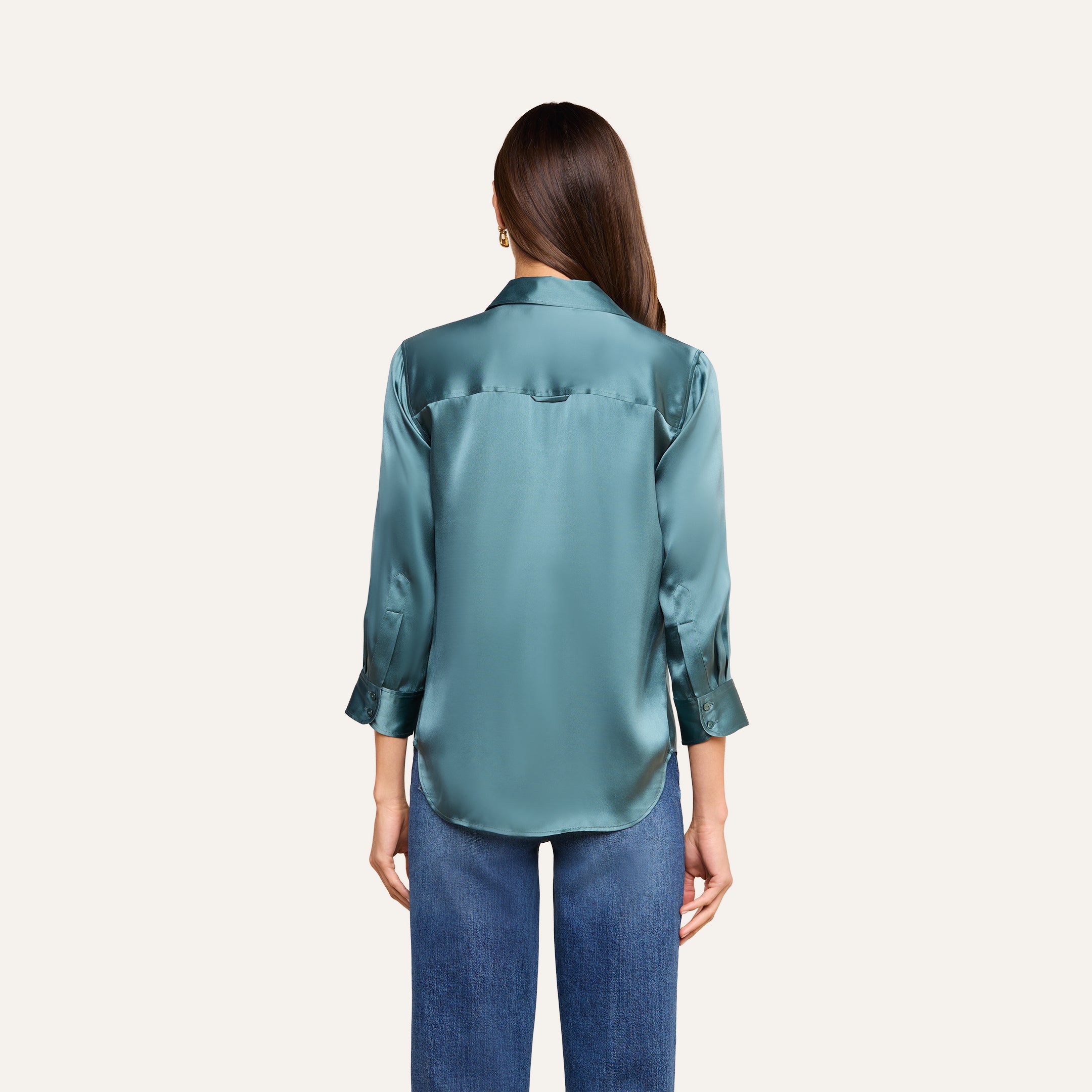 Una mujer con el pelo largo y castaño luce la blusa de seda «Dani» en azul cadete —una lujosa blusa de seda con botones y puños enrollados— combinada con unos vaqueros azules, de pie de espaldas a la cámara sobre un fondo beige.