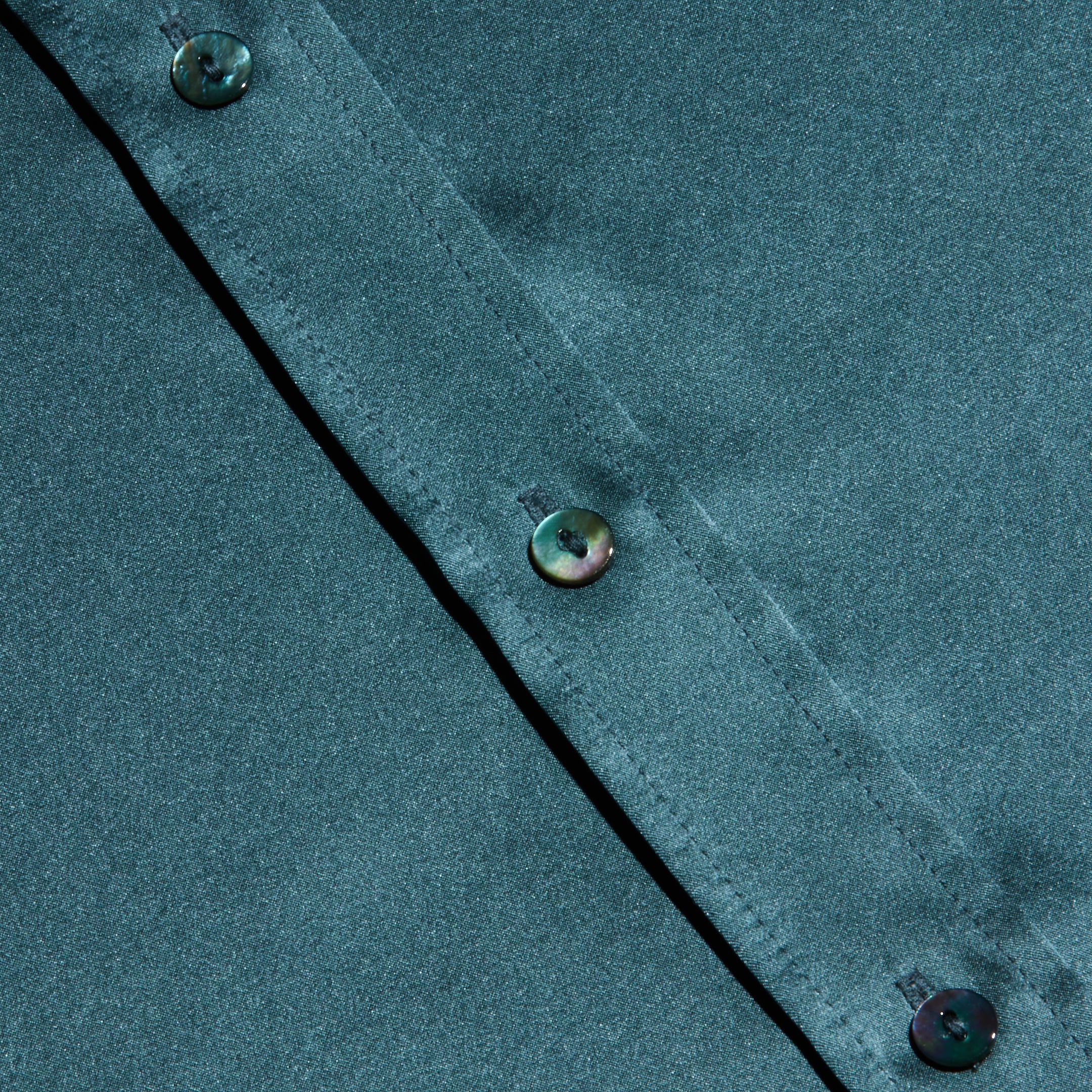 Close-up of a cadet blue Dani Silk Blouse featuring a diagonal row of iridescent buttons. The smooth, slightly shiny silk fabric showcases subtle highlights and shadows across its surface.