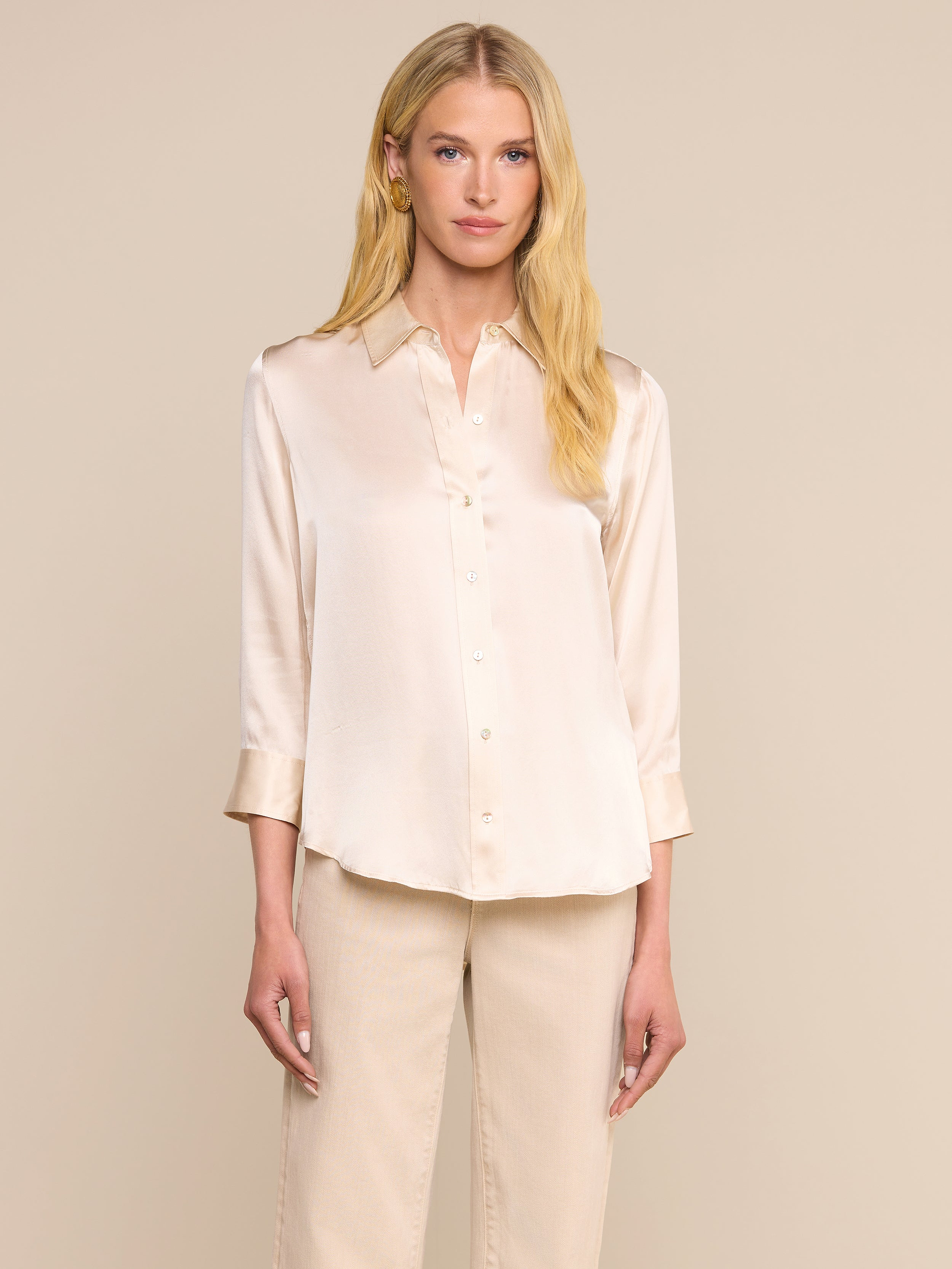 A woman with long blonde hair wears the Dani Silk Blouse in a slim fit champagne silk button-down style, paired with beige pants, standing against a plain light background.