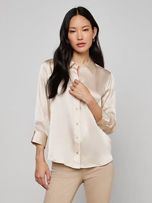 A woman with long black hair stands against a light gray background, wearing the Dani Silk Blouse in shiny champagne satin with beige pants. She has a relaxed expression and touches the collar of her button-down shirt.