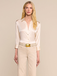A woman with long blonde hair wears the Dani Silk Blouse in champagne, paired with beige pants and a tan belt, standing against an off-white background.