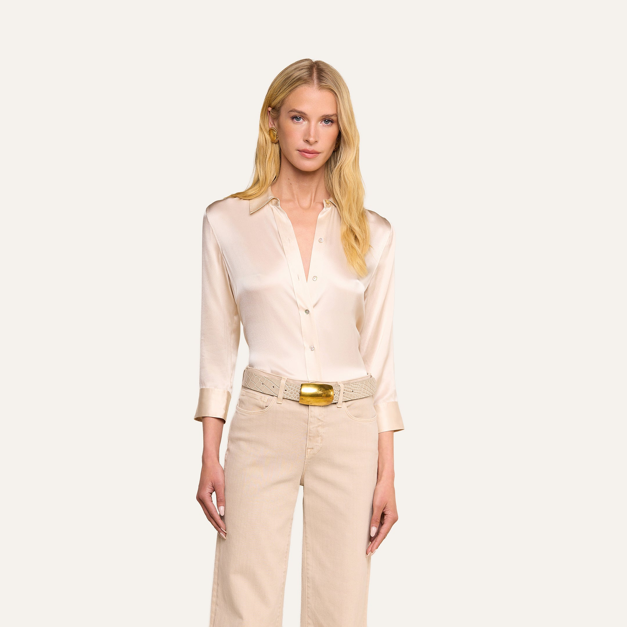 A woman with long blonde hair wears the Dani Silk Blouse in champagne and beige pants with a gold-buckled belt, standing against a plain, light background.