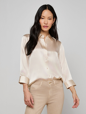 A woman with long black hair wears the Dani Silk Blouse, a slim fit champagne satin button-down with rolled-up sleeves, paired with beige pants. She stands against a plain light gray background.