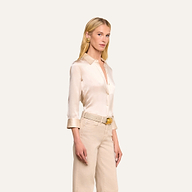 A woman with long blonde hair wears the Dani Silk Blouse in champagne, featuring rolled cuffs, paired with beige high-waisted jeans, a matching belt, and gold hoop earrings, standing against a plain light background.