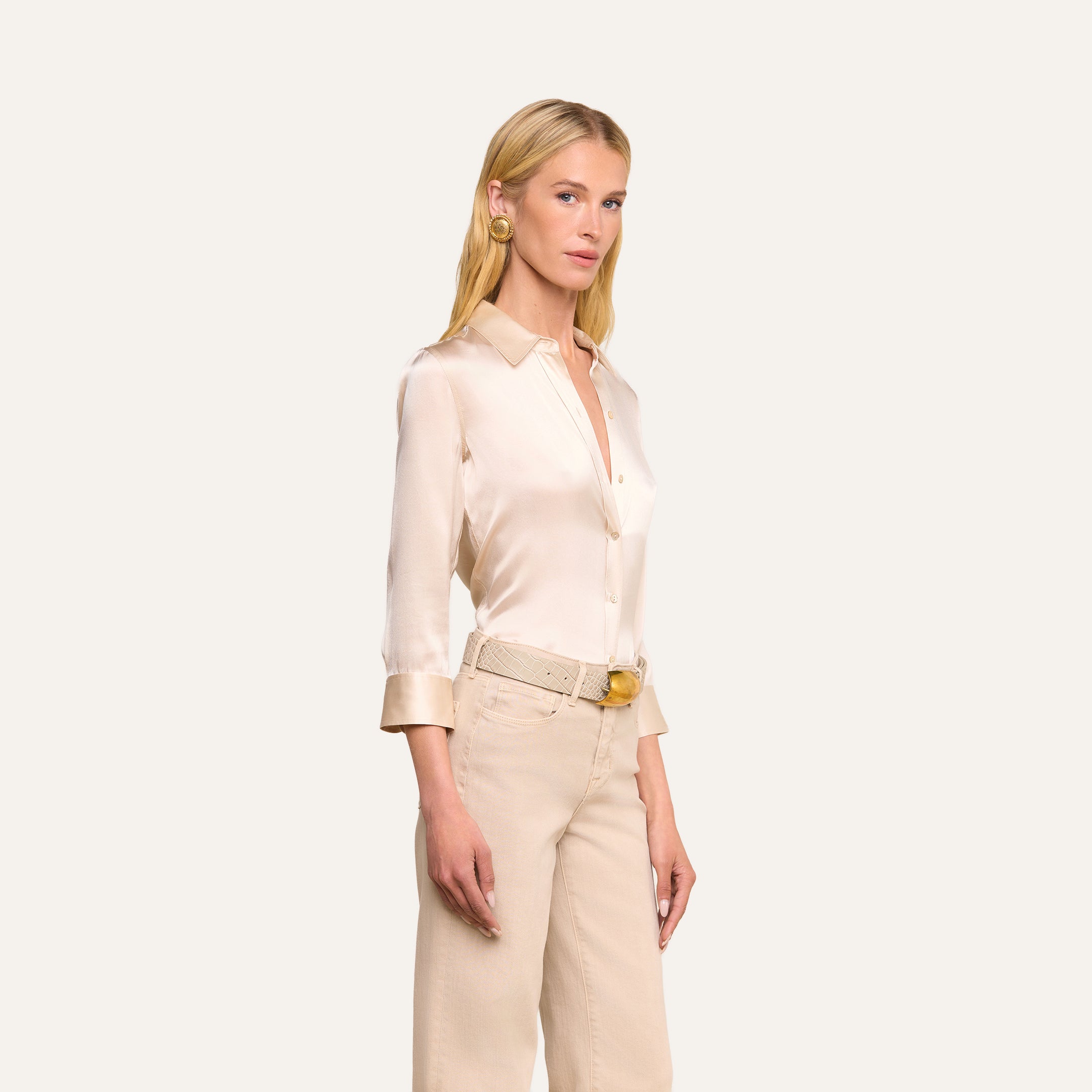 A woman with long blonde hair wears the Dani Silk Blouse in champagne, featuring rolled cuffs, paired with beige high-waisted jeans, a matching belt, and gold hoop earrings, standing against a plain light background.
