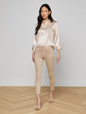 A woman with long dark hair and a neutral expression stands indoors, wearing the Dani Silk Blouse in champagne, paired with fitted beige pants and nude heeled sandals.