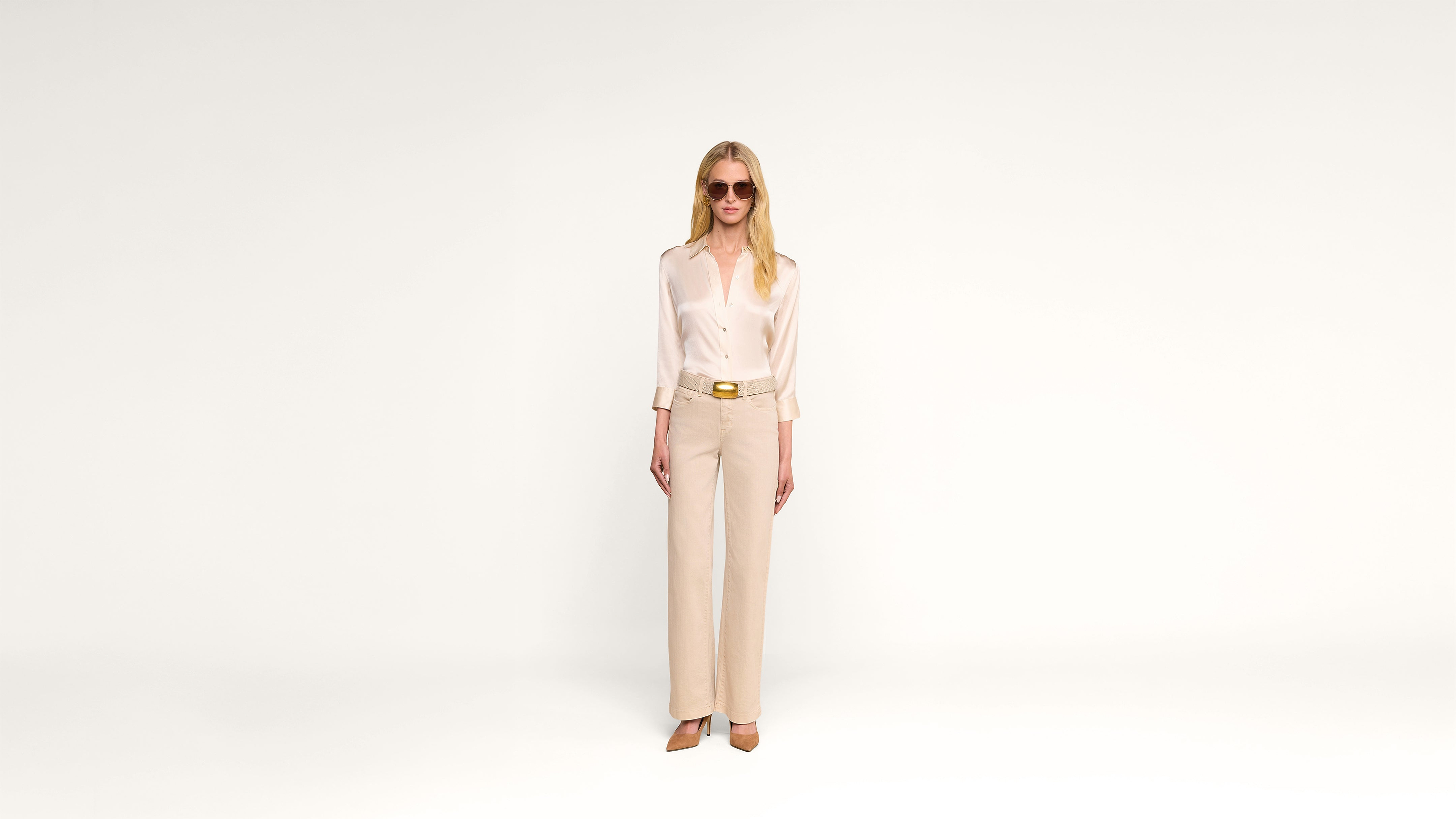 A woman wearing sunglasses, a light silk blouse, beige pants, and heels stands against a plain white background, hands relaxed by her sides.