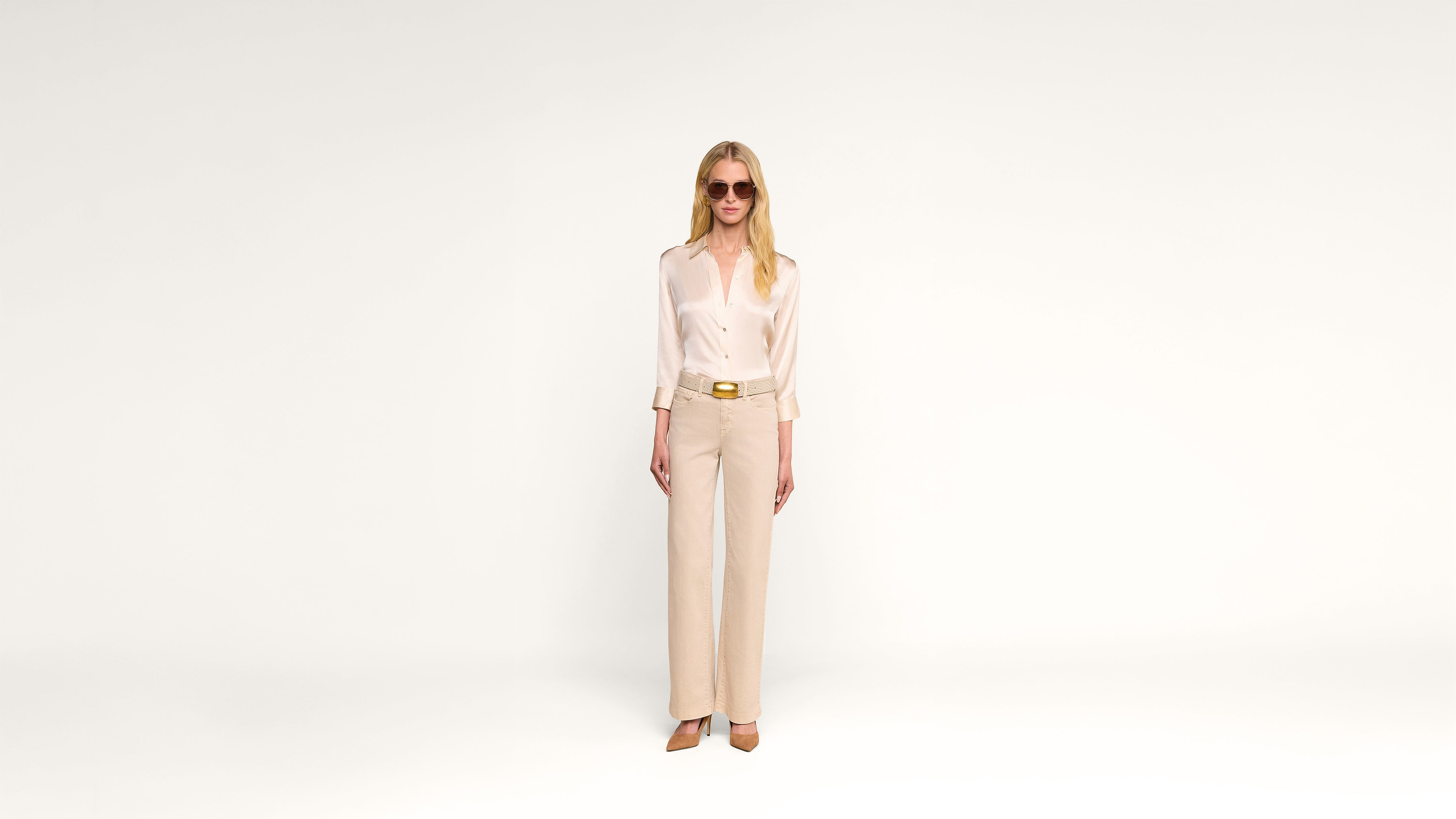 A woman wearing sunglasses, a light silk blouse, beige pants, and heels stands against a plain white background, hands relaxed by her sides.