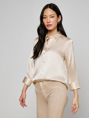 A woman with long dark hair wears the Dani Silk Blouse in champagne, styled with tan pants, posing against a plain light background.