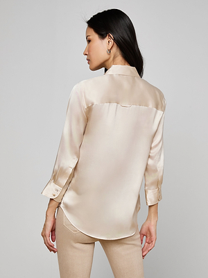 A woman with long dark hair models the Dani Silk Blouse in champagne, paired with beige pants, standing with her back to the camera against a plain light background.