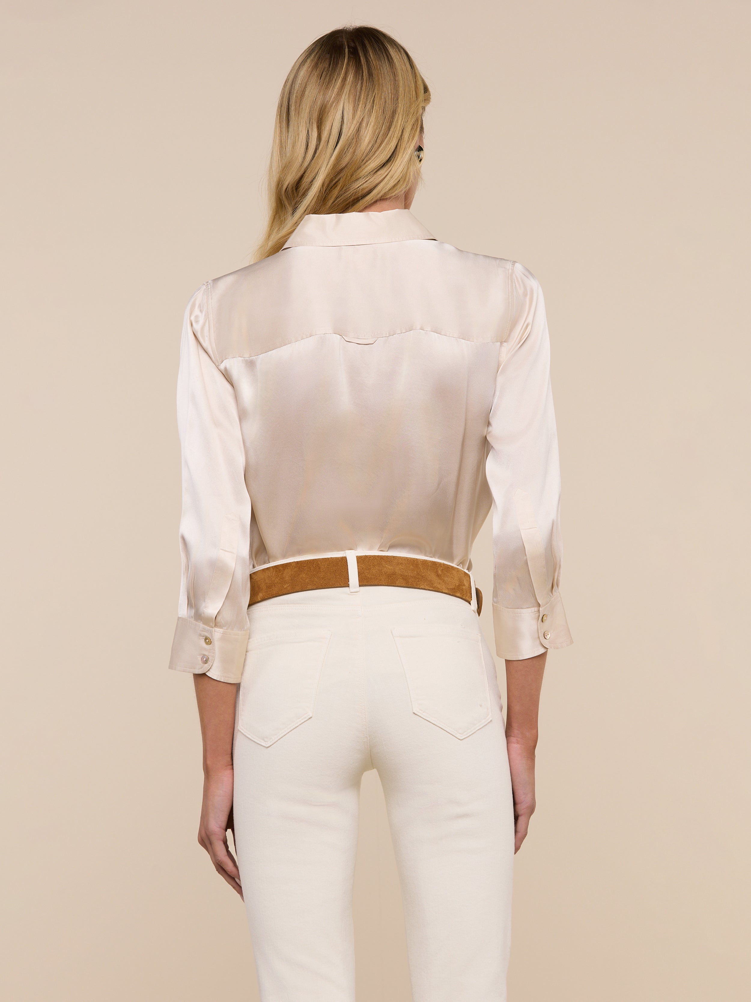 A woman with long blonde hair stands with her back to the camera, wearing the cream-colored Dani Silk Blouse paired with matching pants against a plain light background.