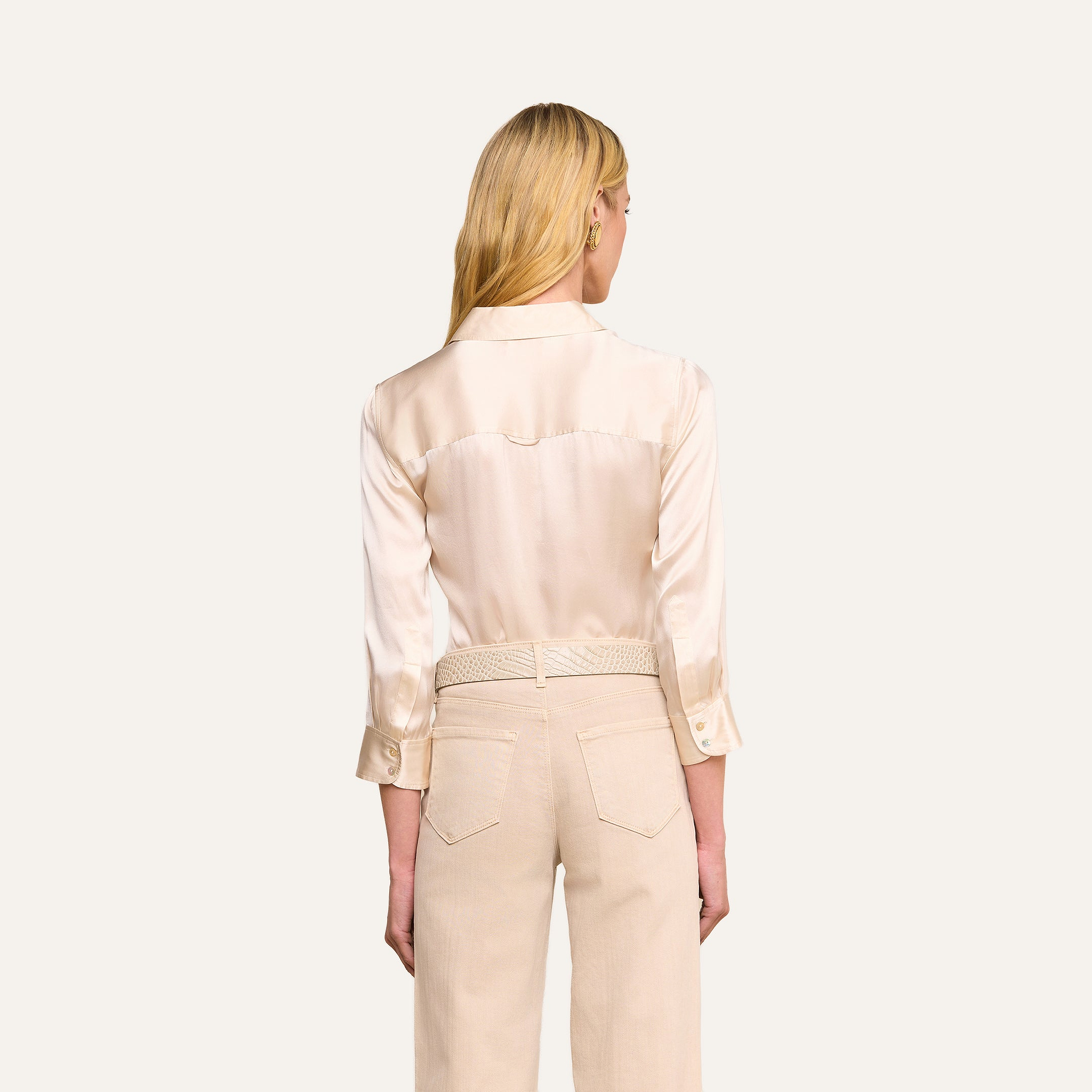 Woman with long blonde hair, shown from the back against a plain light background, wearing the Dani Silk Blouse in champagne paired with light beige high-waisted pants featuring a braided belt.
