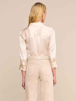 A woman with long blonde hair, seen from the back, wears the cream-colored Dani Silk Blouse with long sleeves and beige slim-fit high-waisted pants, standing against a plain beige background.