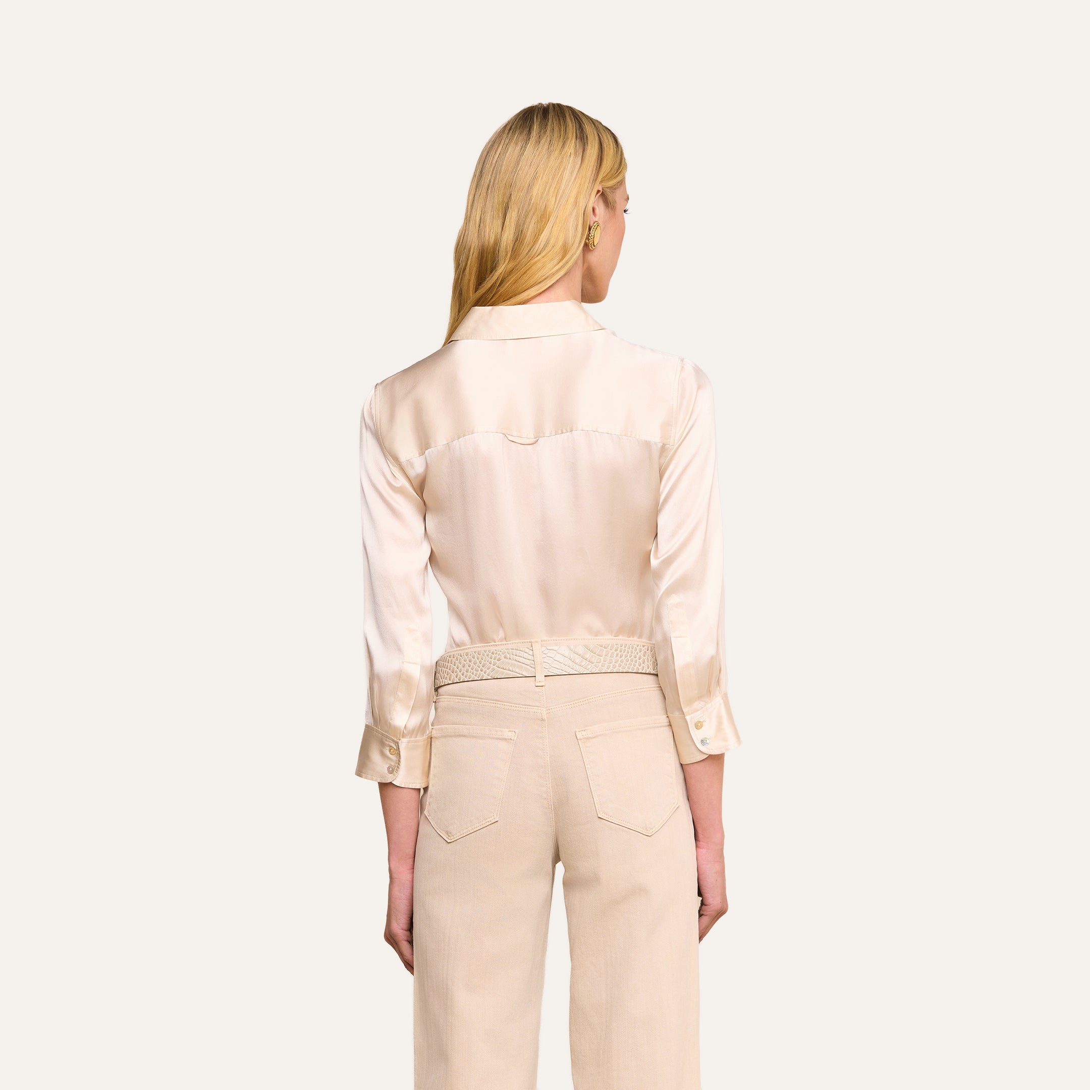 Woman with long blonde hair, shown from the back against a plain light background, wearing the Dani Silk Blouse in champagne paired with light beige high-waisted pants featuring a braided belt.