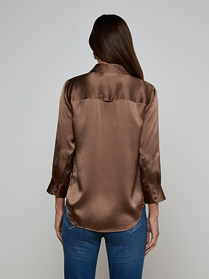 A person with long brown hair is seen from behind, wearing the shiny cardamom brown Dani Silk Blouse with long sleeves and blue jeans against a plain background.