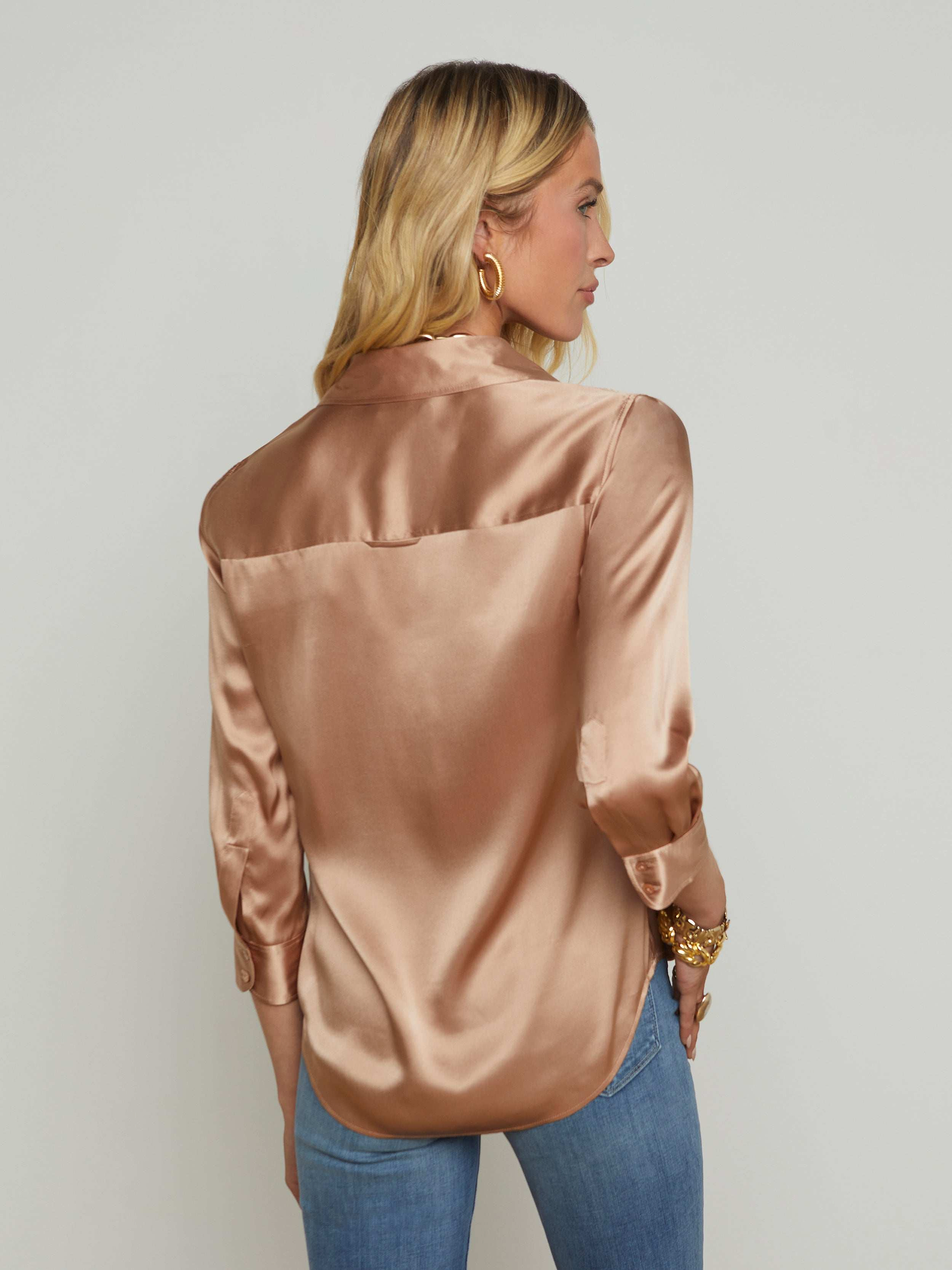 A woman with blonde hair, wearing gold hoop earrings, the Dani Silk Blouse in a shiny light brown hue, and blue jeans stands with her back to the camera against a plain light background.