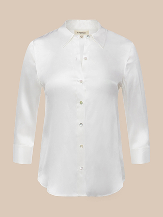 La blusa Dani Silk es una prenda blanca de manga larga confeccionada en seda charmeuse, con un cuello clásico y un acabado sutilmente brillante, que destaca sobre un fondo beige liso.