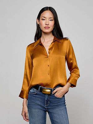 A woman with long dark hair wears the L'AGENCE Dani Silk Blouse—a shiny copper button-down with three-quarter sleeves—paired with blue jeans and a wide belt, standing against a plain gray backdrop.