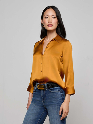 A woman with long dark hair wears the L'AGENCE Dani Silk Blouse in shiny gold, featuring three-quarter sleeves and a button-down design, paired with blue jeans against a light gray background, looking slightly to the side.