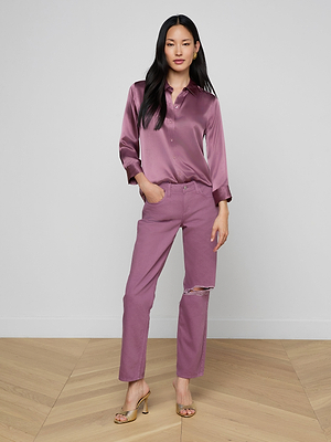 A woman with long, dark hair stands confidently indoors on a wooden floor, wearing the Dani Silk Blouse in mauve, matching mauve pants with a knee rip, and gold open-toe heels.