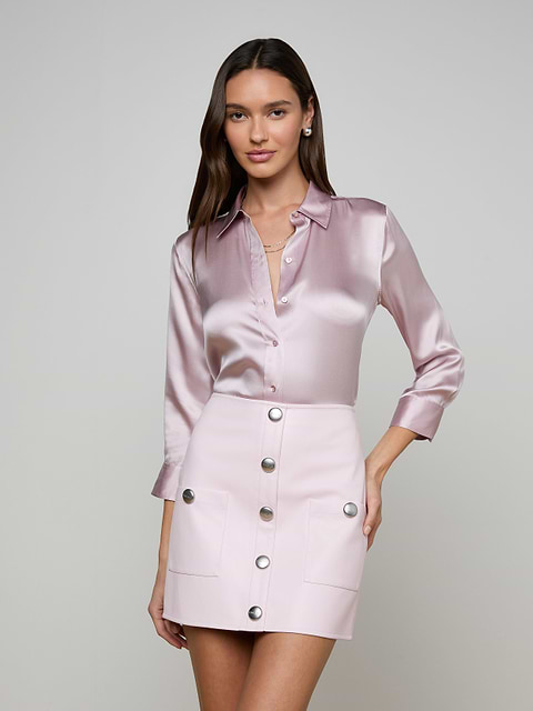 A woman stands against a plain off-white background, wearing a shiny light pink Dani Silk Blouse with rolled sleeves paired with a matching light pink mini skirt adorned with large silver buttons down the front.