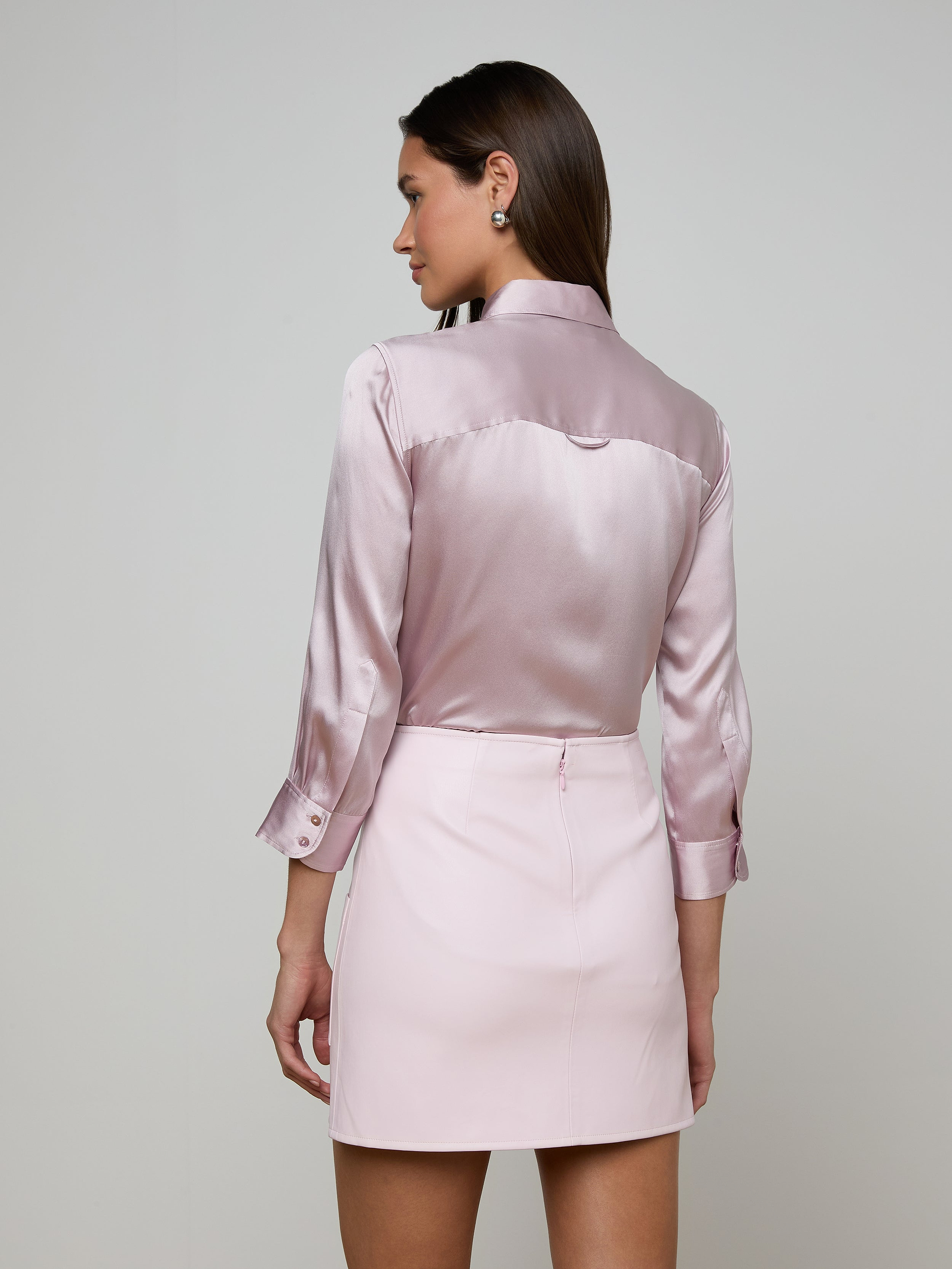 A woman with straight brown hair is shown from behind, wearing a shiny, light pink Dani Silk Blouse with long sleeves and a matching mini skirt. She stands against a plain light background.