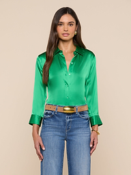A woman with long brown hair wears the Dani Silk Blouse—shiny green with 3/4 sleeves, wide cuffs, and button-down front—styled with blue jeans, a tan belt featuring a gold buckle, and gold jewelry. She stands against a plain light background.