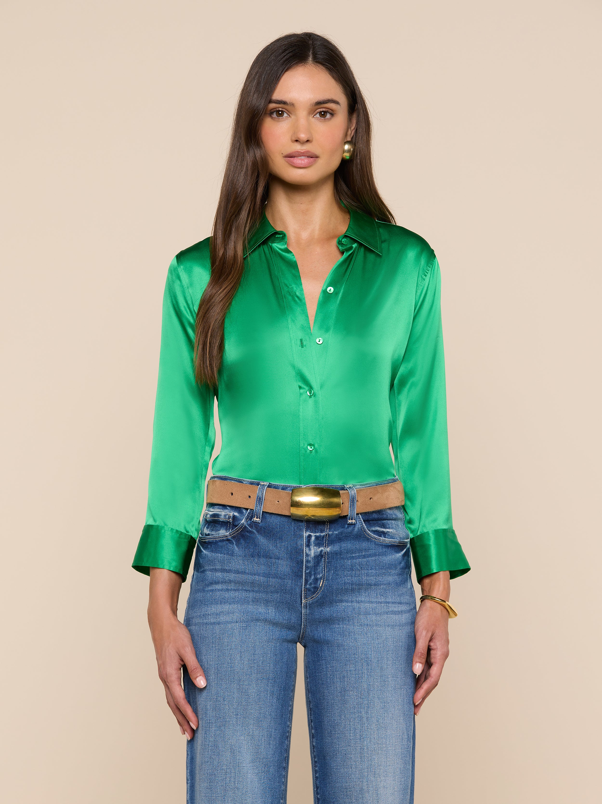 A woman with long brown hair wears the Dani Silk Blouse—shiny green with 3/4 sleeves, wide cuffs, and button-down front—styled with blue jeans, a tan belt featuring a gold buckle, and gold jewelry. She stands against a plain light background.