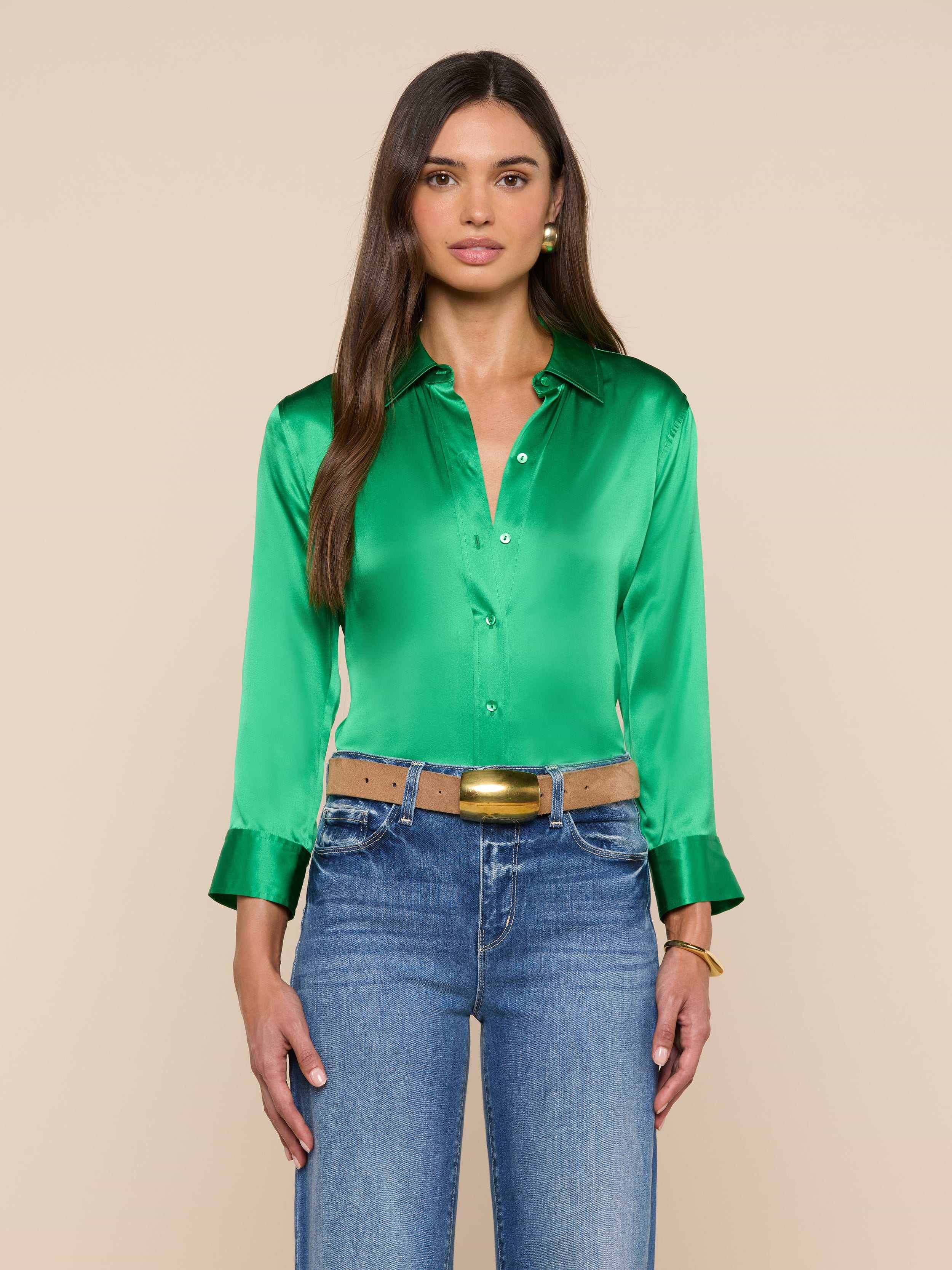 A woman with long brown hair wears the Dani Silk Blouse—shiny green with 3/4 sleeves, wide cuffs, and button-down front—styled with blue jeans, a tan belt featuring a gold buckle, and gold jewelry. She stands against a plain light background.