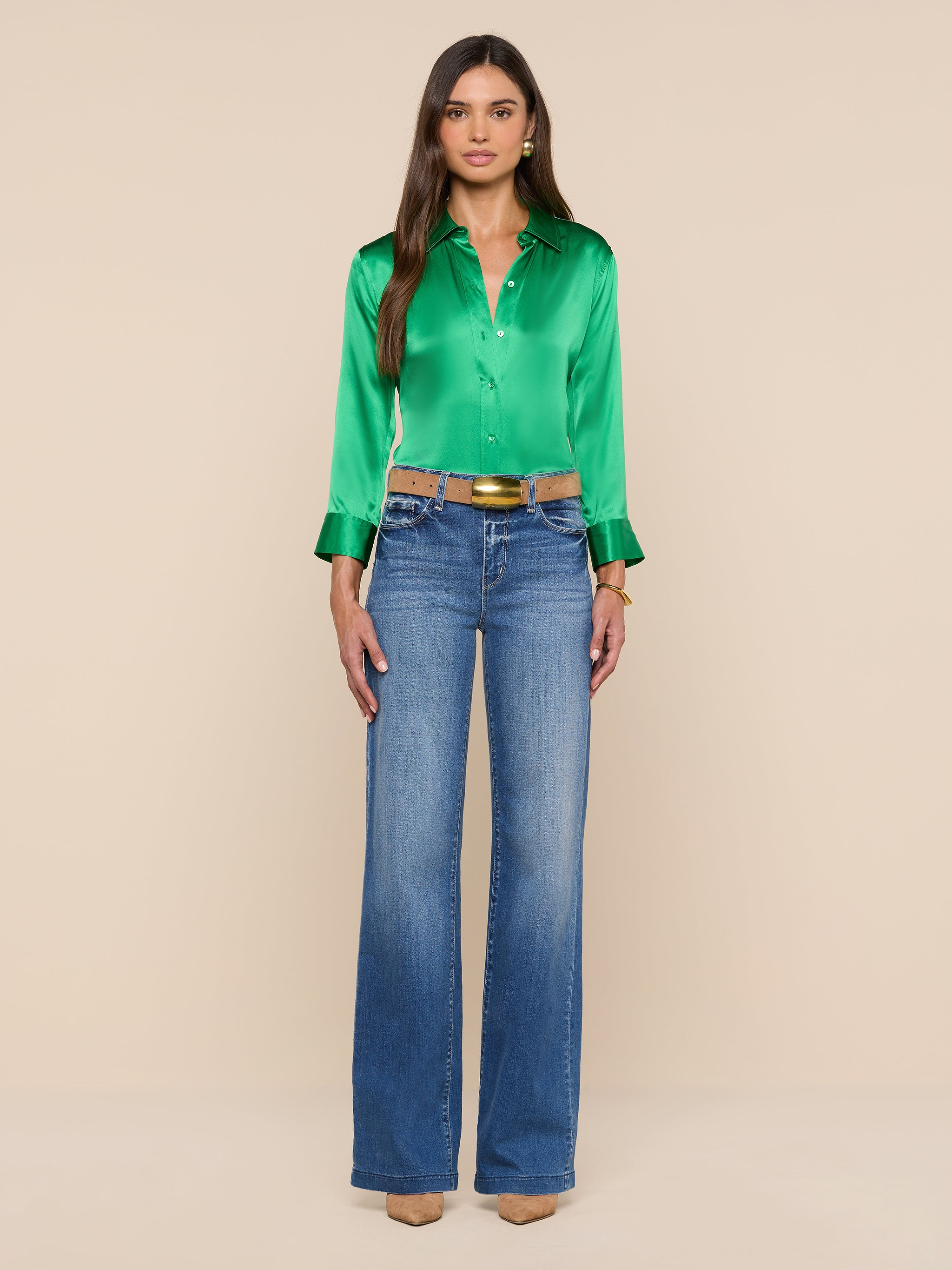 A woman stands facing forward, wearing a shiny green Dani Silk Blouse with 3/4 sleeves, blue high-waisted wide-leg jeans, and tan heels. The background is plain white.