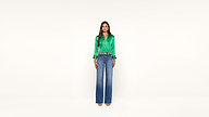 A woman stands facing forward, wearing a bright green satin blouse, blue high-waisted wide-leg jeans, and beige heels against a plain white background.