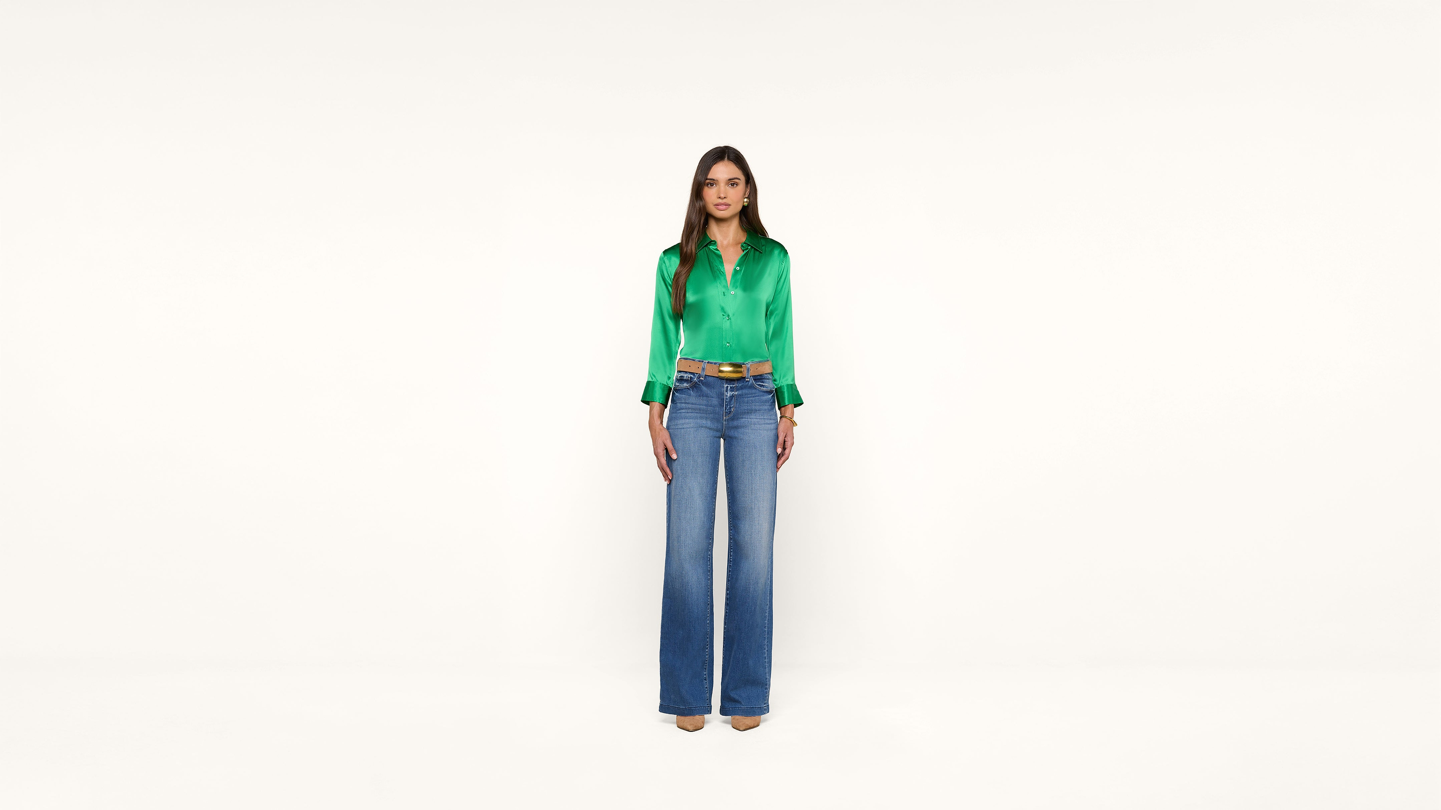 A woman stands facing forward, wearing a bright green satin blouse, blue high-waisted wide-leg jeans, and beige heels against a plain white background.