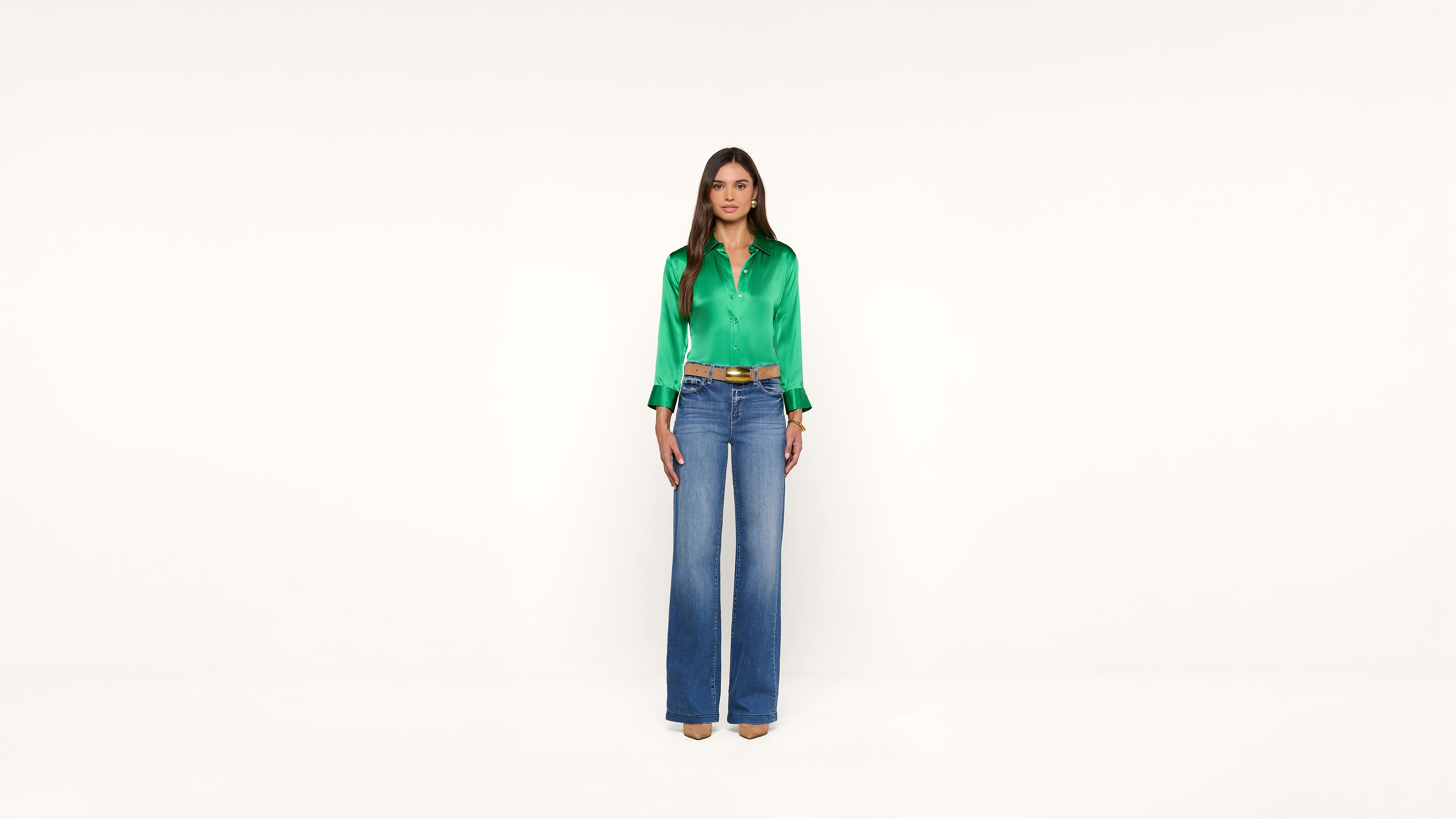 A woman stands facing forward, wearing a bright green satin blouse, blue high-waisted wide-leg jeans, and beige heels against a plain white background.