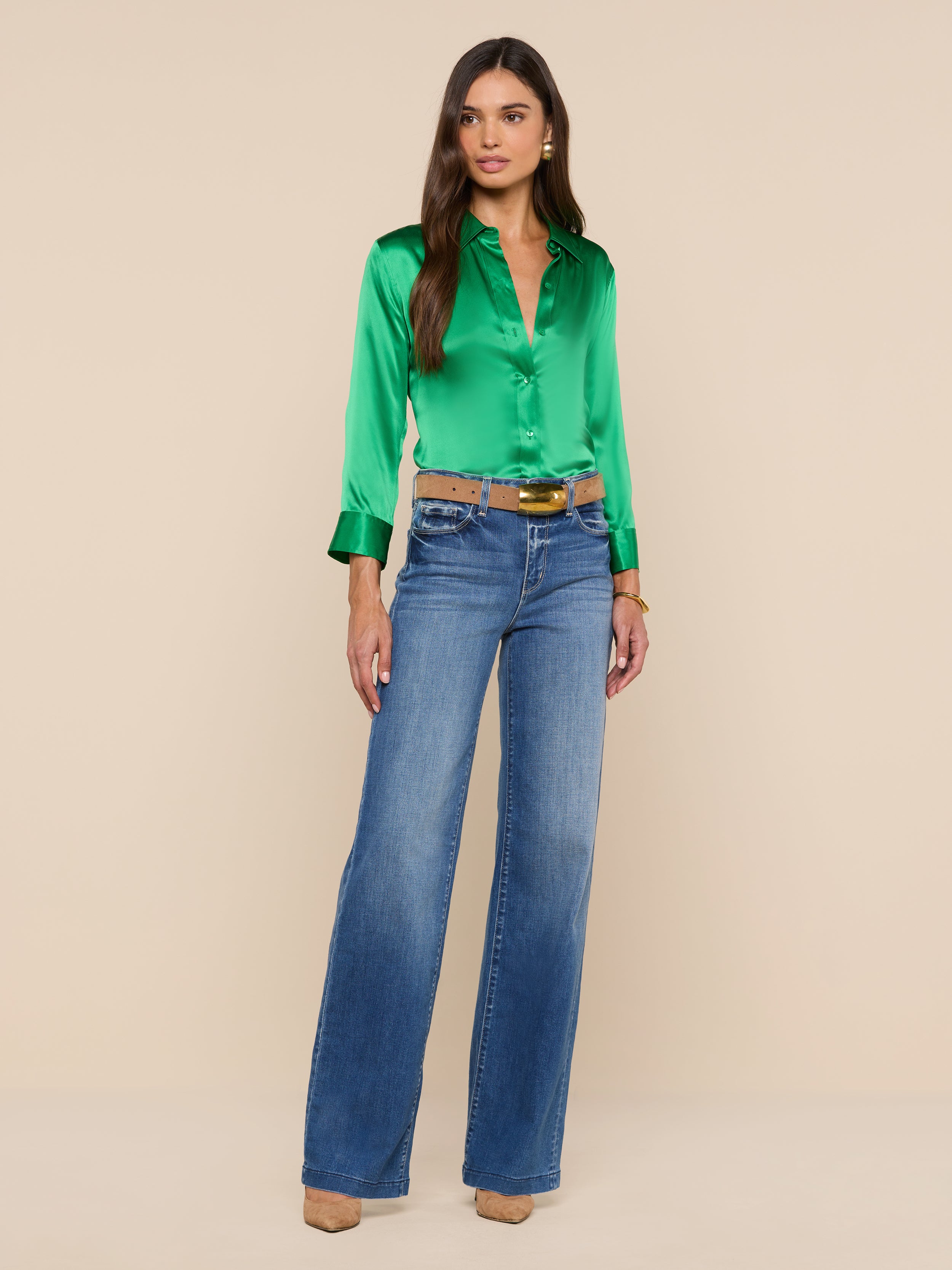 A woman stands against a plain white background, wearing a shiny green Dani Silk Blouse with 3/4 sleeves, blue high-waisted wide-leg jeans, a brown belt, and tan heels. She has long brown hair and a neutral expression.