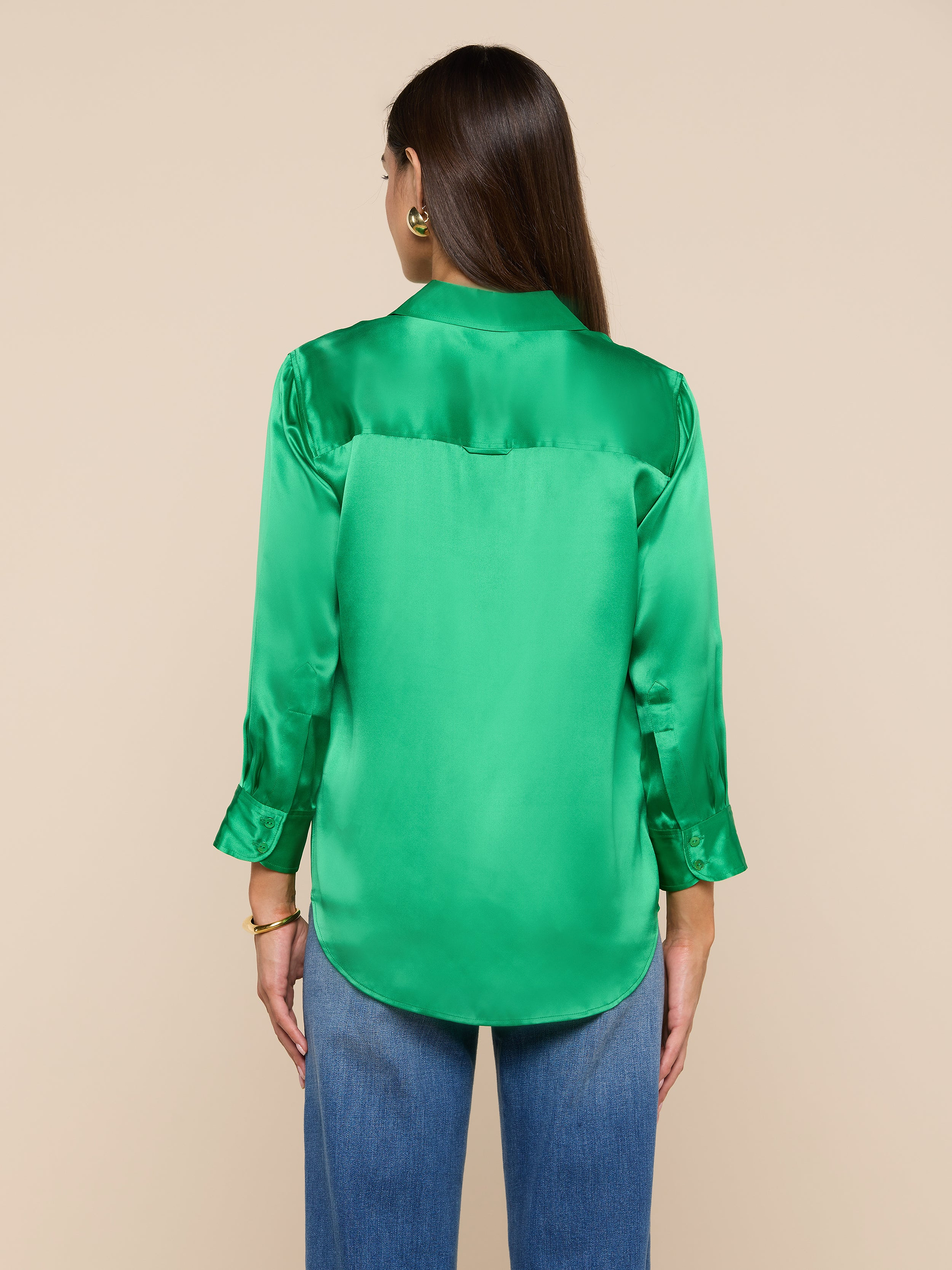 A woman with long brown hair is shown from the back, wearing a bright green Dani Silk Blouse with long sleeves and blue jeans, standing against a plain light background.