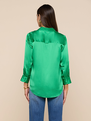 A woman with long brown hair is seen from behind, wearing the bright green Dani Silk Blouse with 3/4 sleeves and blue jeans, standing against a plain beige background.