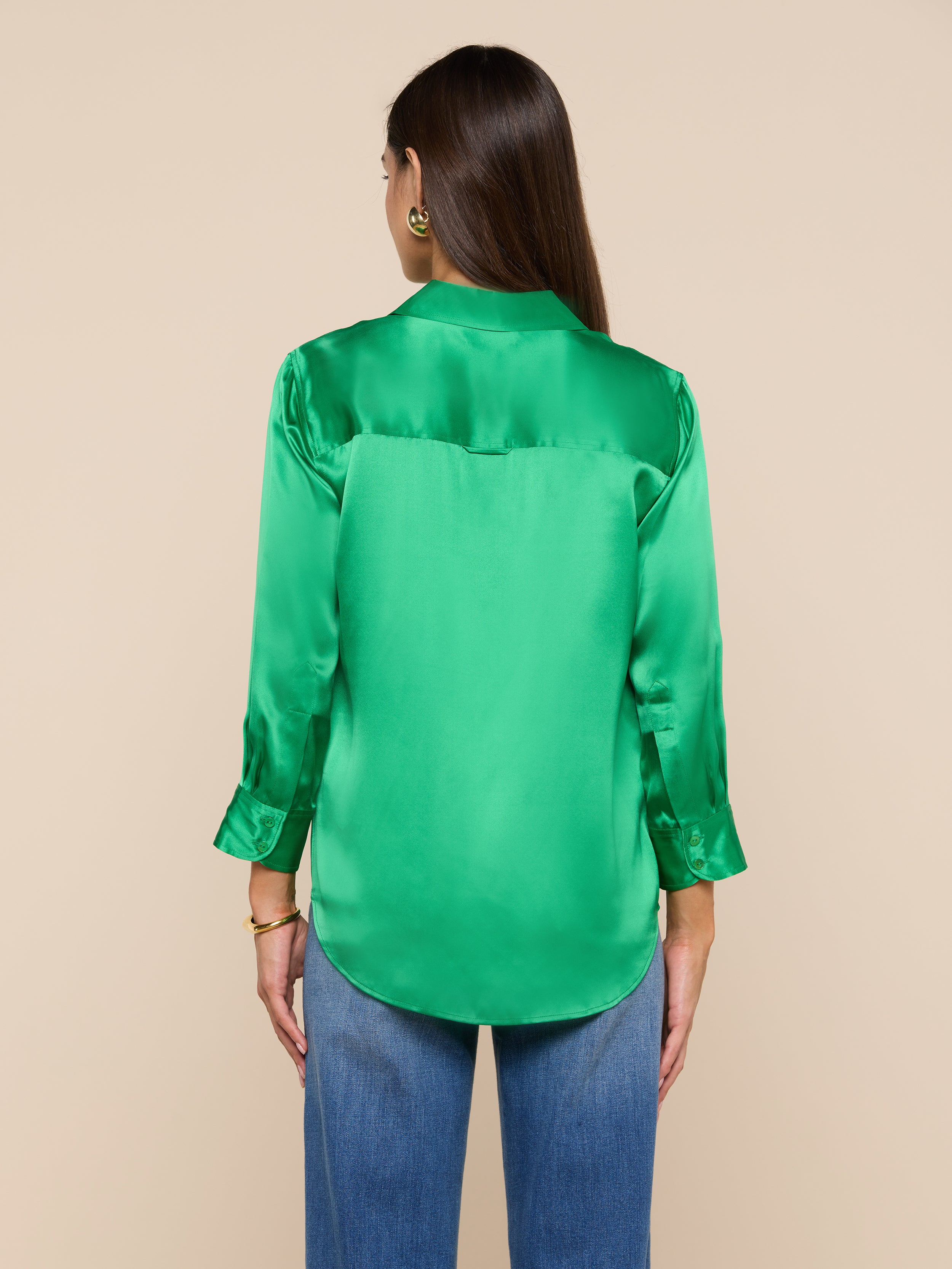 A woman with long brown hair is shown from the back, wearing a bright green Dani Silk Blouse with long sleeves and blue jeans, standing against a plain light background.