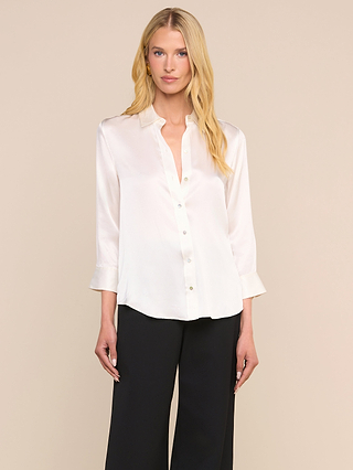 Una persona con el pelo largo y rubio lleva la blusa Dani Silk, una prenda de color marfil con botones y mangas tres cuartos, combinada con unos pantalones negros de corte ancho, de pie frente a un fondo liso de color beige.