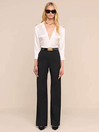 Una mujer con gafas de sol y la blusa «Dani Silk», metida por dentro de unos pantalones negros de cintura alta con un cinturón ancho y combinada con unos zapatos de tacón negros, posa frente a un fondo beige.