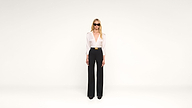 A woman wearing sunglasses, a white blouse, and high-waisted black pants stands against a plain white background.